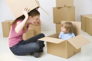 A woman with young girl sitting between cardboard boxes. Happy woman holding box over head and looking at young girl. Young girl sitting in box.