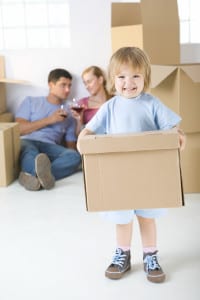 Young parents sitting beside cardboard boxes and drinking wine. Theirs daughter standing in frond and holding box. She's looking at camera.